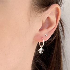 May include: Silver hoop earrings with a delicate flower charm hanging from the bottom. The earrings are worn in a person's ear.