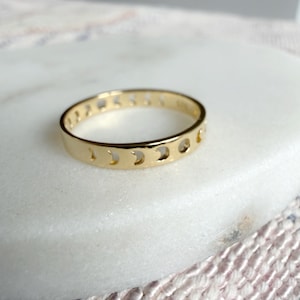 May include: A gold ring with a repeating crescent moon design. The ring is displayed on a white marble surface. The ring's design is consistent around the entire band. The ring is circular.