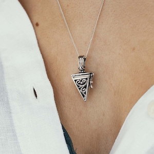 Puede incluir: Un collar con colgante de plata con un colgante triangular y ornamentado. El colgante presenta intrincados adornos y un diseño con bisagras. El collar se lleva sobre una camisa blanca.
