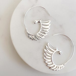 Puede incluir: Un par de pendientes plateados en espiral con un diseño en forma de ala. Los pendientes están sobre una superficie de mármol blanco. El diseño del ala presenta múltiples líneas paralelas, creando una apariencia de pluma. Joyería moderna.