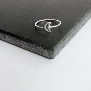 May include: A silver-colored ring featuring a crescent moon design. The ring is set against a dark gray, textured surface, with a white background. The ring's band is thin and delicate, and the moon shape is open.