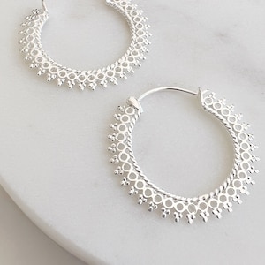 May include: A pair of silver hoop earrings. The earrings feature a detailed design with small circular cutouts and beaded edges. The hoops are displayed on a white surface, highlighting their intricate craftsmanship and elegant style.