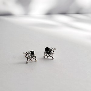 May include: Two silver-toned stud earrings. Each earring features a black, circular stone at the center, surrounded by a stylized floral design. The earrings are set against a white background.