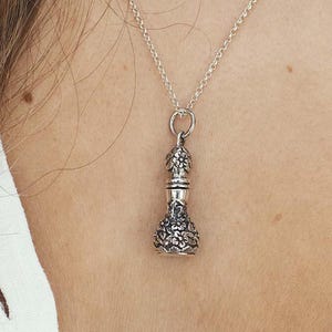 May include: A silver pendant necklace featuring a detailed, floral-patterned charm. The charm has a cylindrical shape with a decorative top and bottom. The necklace chain is delicate and silver, creating an elegant accessory.