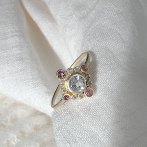 Puede incluir: Un anillo de oro con una piedra blanca rodeada de piedras blancas y rosas más pequeñas.