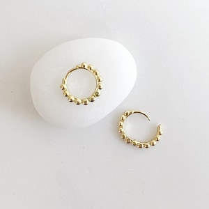 Op de afbeelding: Twee goudkleurige oorringen met een kralenontwerp, gepresenteerd op een witte, gladde ondergrond. De oorbellen zijn klein en cirkelvormig, met een reeks kleine, ronde kralen die de ringen omlijnen. De oorbellen zijn een sieraad.