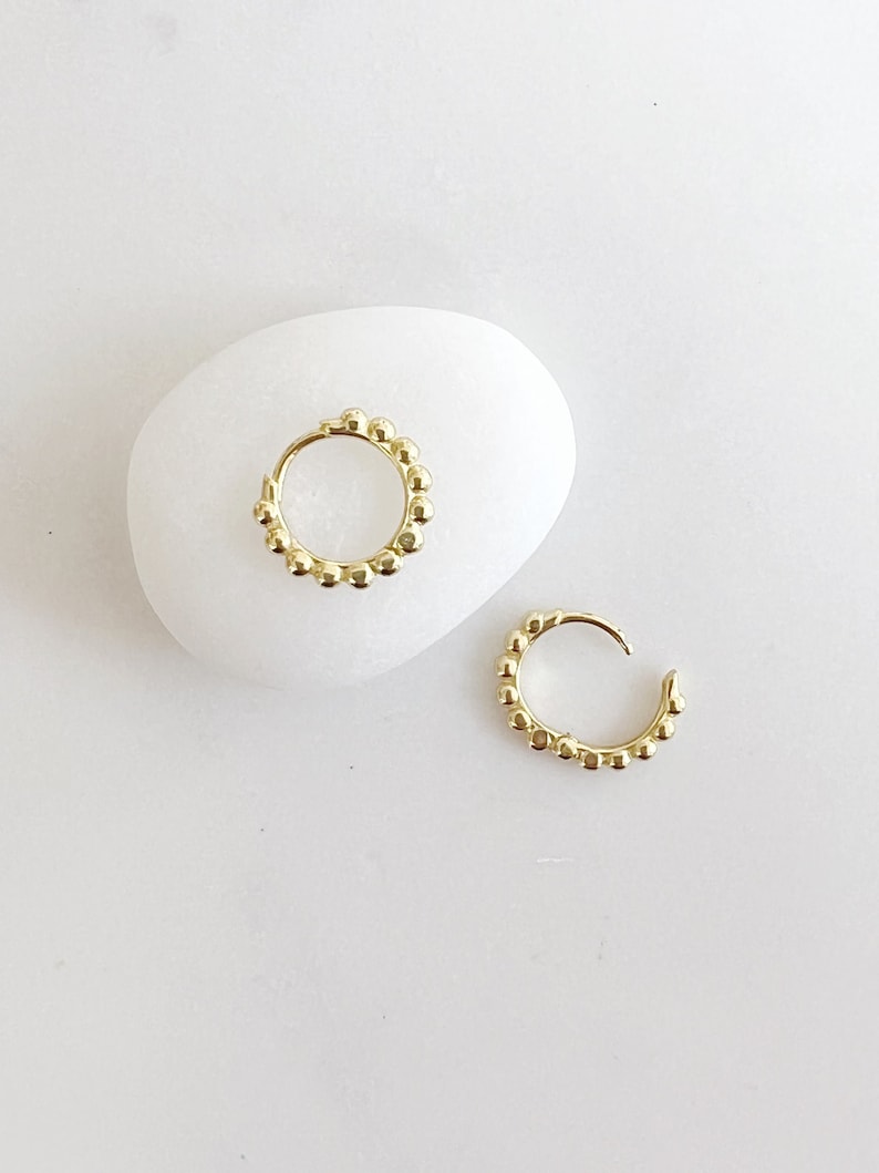 Op de afbeelding: Twee goudkleurige oorringen met een kralenontwerp, gepresenteerd op een witte, gladde ondergrond. De oorbellen zijn klein en cirkelvormig, met een reeks kleine, ronde kralen die de ringen omlijnen. De oorbellen zijn een sieraad.