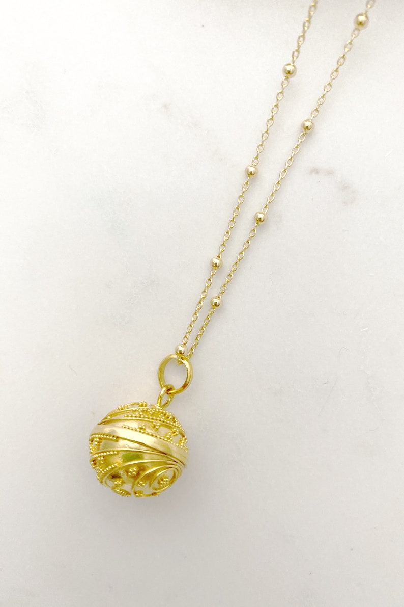 May include: A gold necklace with a delicate chain and a round, ornate pendant. The pendant has a detailed, intricate design and appears to be hollow.