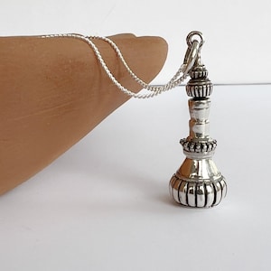 Può includere: Una collana con pendente color argento con un design ornato e dettagliato. Il pendente ha una base bulbosa e un corpo cilindrico, con un piccolo anello per la catena. La catena è in argento intrecciato delicato.