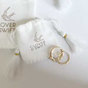 Op de afbeelding: Witte zakjes met trekkoord met gouden tekst en een geometrisch vogel logo, en gouden oorringen. De zakjes hebben witte kwastjes met gouden accenten. De tekst op het zakje luidt "CLOVER & SWIFT".