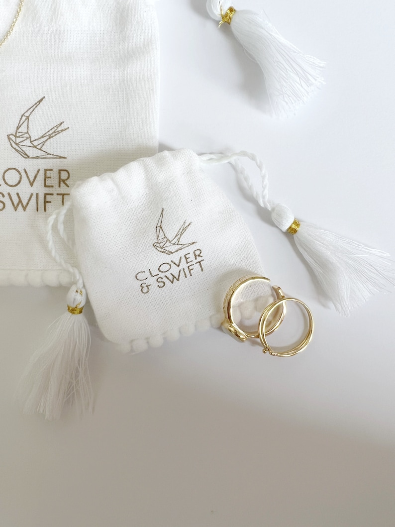 Op de afbeelding: Witte zakjes met trekkoord met gouden tekst en een geometrisch vogel logo, en gouden oorringen. De zakjes hebben witte kwastjes met gouden accenten. De tekst op het zakje luidt "CLOVER & SWIFT".