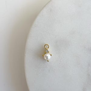 May include: Gold charm with a small pearl and a clear rhinestone. The charm is designed to be added to a necklace or bracelet.