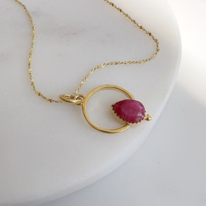 Op de afbeelding: Een goudkleurige ketting met een hanger met een druppelvormige, dieprode edelsteen. De edelsteen is gezet in een goudkleurige bezel en een goudkleurige cirkelvormige lijst. De fijne ketting heeft een subtiele glans.