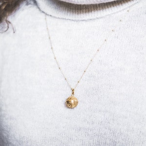 May include: A gold necklace with a small, ornate, round pendant hangs on a white sweater.