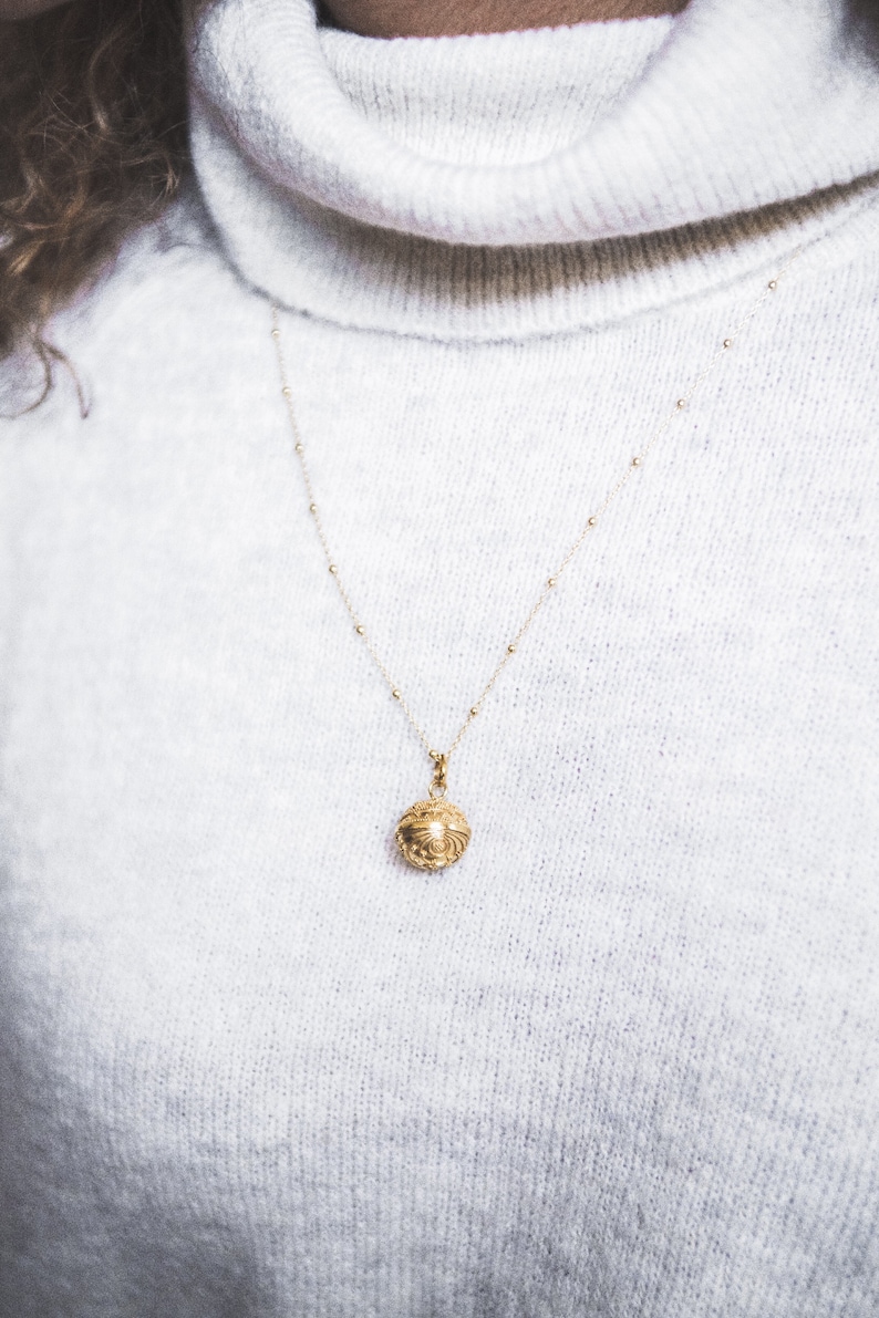 May include: A gold necklace with a small, ornate, round pendant hangs on a white sweater.