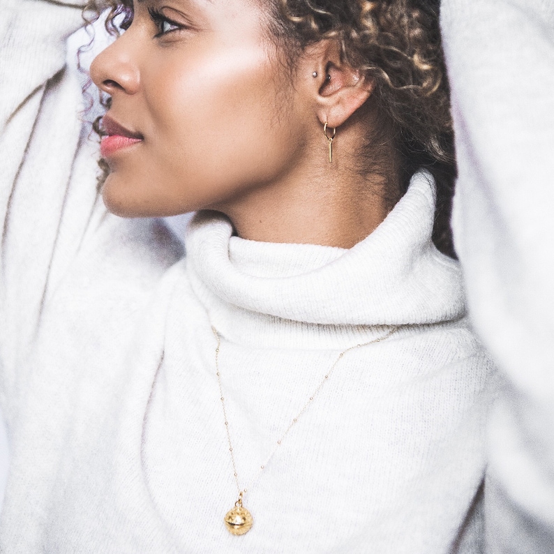May include: A woman wearing a white turtleneck sweater with a gold necklace featuring a small, round pendant. The necklace is delicate and has a simple design.