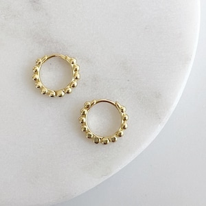 Op de afbeelding: Twee goudkleurige oorringen. Elke oorring is versierd met kleine, gelijkmatig verdeelde, bolvormige kralen rond de ring. De oorbellen worden gepresenteerd op een wit marmeren oppervlak.
