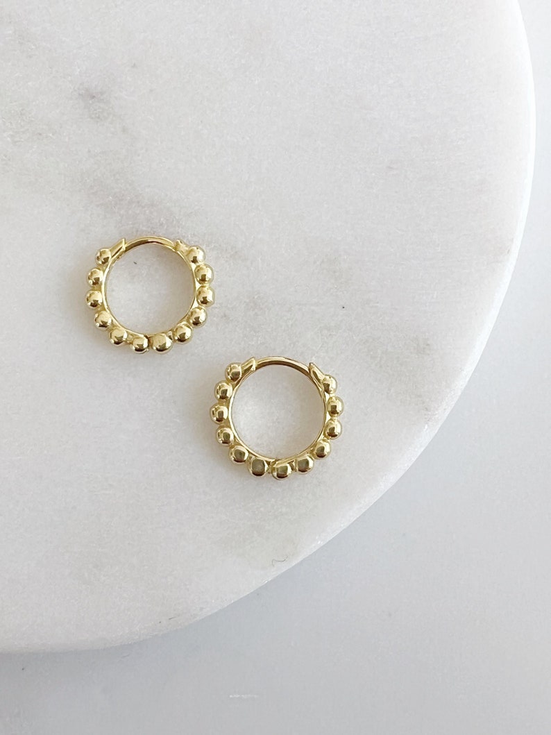 Op de afbeelding: Twee goudkleurige oorringen. Elke oorring is versierd met kleine, gelijkmatig verdeelde, bolvormige kralen rond de ring. De oorbellen worden gepresenteerd op een wit marmeren oppervlak.