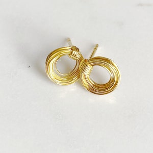 Può includere: Un paio di orecchini a bottone color oro. Ogni orecchino presenta un design circolare realizzato con filo avvolto. Gli orecchini sono su una superficie bianca. Gli orecchini sono piccoli e delicati.