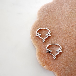 May include: A pair of silver hoop earrings featuring a heart-shaped design and small beaded details. The earrings are presented on a textured, light brown surface. The earrings are a jewelry item, showcasing their intricate design.
