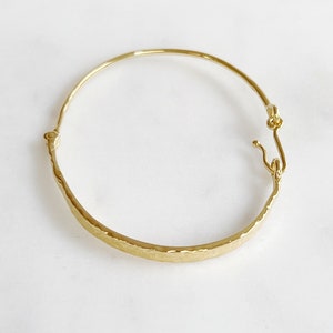 Peut inclure: Un bracelet jonc de couleur or avec une texture martelée. Le bracelet a un design simple et élégant avec une fermeture à crochet et œillet. Le bracelet est présenté sur une surface blanche.