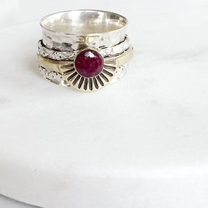 Peut inclure: Une bague rotative argentée et dorée avec une grosse pierre rouge foncé et ronde. La bague a une texture martelée sur la bande supérieure et un motif de soleil autour de la pierre précieuse. La bague est posée sur une surface blanche.