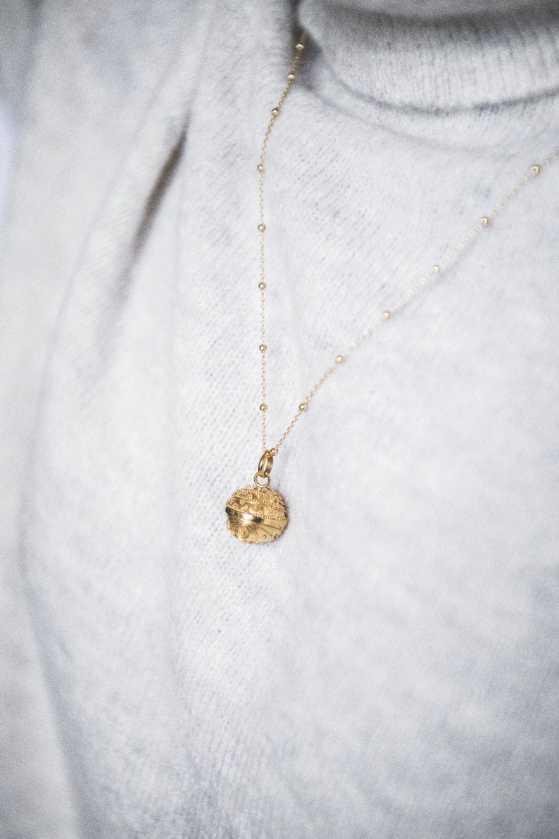 May include: A gold necklace with a round, textured pendant. The necklace is on a white fuzzy sweater.