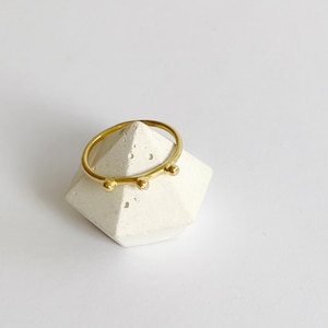 Puede incluir: Un anillo de oro con pequeños acentos redondos en un soporte de anillo geométrico blanco.