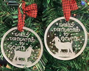 May include: Two round glass ornaments with red and black checkered ribbon. The ornaments are white with a goat silhouette and the text "Easily Distracted by Goats".