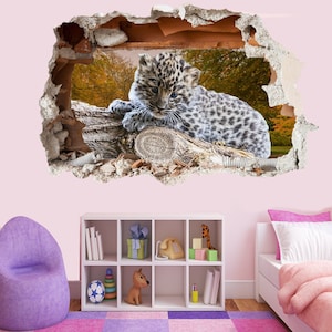 Puede incluir: Un cachorro de leopardo blanco y gris yace sobre una rama de árbol, mirando a la cámara. El cachorro está en una pared rota, creando un efecto 3D. El fondo es una imagen borrosa de un bosque.