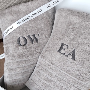 Op de afbeelding: Twee grijze handdoeken met geborduurde initialen. De handdoek links heeft de initialen "OW" geborduurd. De handdoek rechts heeft de initialen "EA" geborduurd. De handdoeken zijn samen verpakt met een wit lint waarop "The Stitch Company" staat.