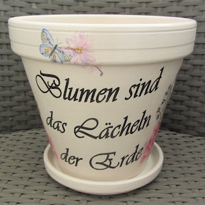 Könnte beinhalten: Weißer Keramik-Blumentopf mit einem gemalten Schmetterling und Blumen. Der Topf hat eine schwarze Aufschrift mit dem Text "Blumen sind das Lächeln der Erde".