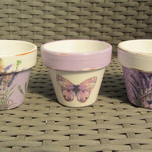 Könnte beinhalten: Drei kleine Terrakotta-Blumentöpfe mit Lavendel-Designs. Die Töpfe sind in Weiß-, Lila- und Grüntönen bemalt. Ein Topf zeigt einen Schmetterling, ein anderer ein Vogelhaus und der dritte einen Lavendelstrauß.
