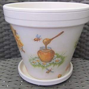 Op de afbeelding: Witte keramische bloempot met een schotel. De pot is versierd met een honingbij- en honingpotontwerp. Het ontwerp bevat bijen, honing, madeliefjes en een houten lepel. De pot staat op een grijze rieten stoel.