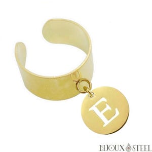 Peut inclure: Une bague ajustable dorée avec un charm circulaire. Le charm présente la lettre "E" découpée au centre. La bague est en métal brillant sur fond blanc.