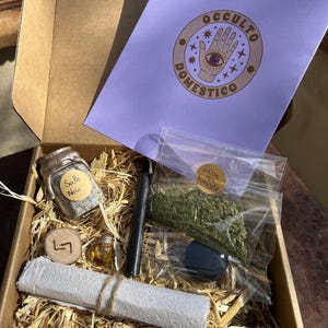 Può includere: Una scatola di cartone riempita di paglia contenente un piccolo barattolo di vetro con etichetta Sale Nero, un bastoncino nero, una piccola fiala, un disco di legno con un simbolo, un foglio di carta bianco arrotolato e un sacchetto di plastica con erbe verdi, oltre a un cartoncino viola con il testo Occulto Domestico.
