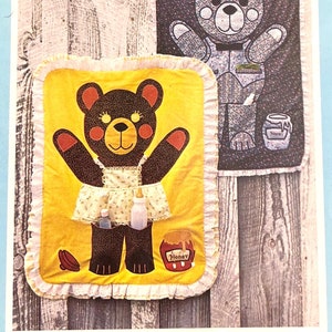 May include: Two quilted wall hangings featuring a bear design. The first shows a gray bear wearing a blue and white patterned shirt and holding a jar of honey. The second shows a brown bear wearing a yellow dress and holding a bottle and a jar of honey. Both hangings have a white ruffled border.