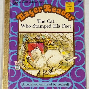 May include: A vintage children's book titled "Eager Reader: The Cat Who Stamped His Feet." The cover features a white cat in a room, with text in a brown and orange font. The book has a purple and blue patterned border.