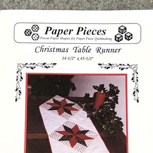 May include: A Christmas table runner pattern with a white background and red, black, and white stars. The pattern includes precut paper pieces for 3-inch 8-point diamonds, squares, and half squares. The table runner measures 14-1/2 inches by 43-1/2 inches.