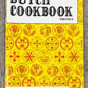 May include: A vintage Dutch Cookbook, Volume II, with a bright yellow cover. The cover features the title "DUTCH COOKBOOK" in bold black letters, and is decorated with red and yellow floral and geometric patterns. The author's name, Edna Eby Heller, is also displayed.