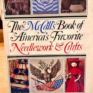 May include: A white hardcover book titled The McCall's Book of America's Favorite Needlework & Crafts. The cover features six rectangular color photographs displaying various craft projects, including a fireplace, a shell pattern, and a doll.