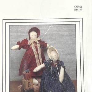 May include: Two handmade cloth dolls, one in a red patterned dress and the other in a blue floral dress. The dolls have white faces and are wearing bonnets. Wooden alphabet blocks with the letters B and C are in front of the dolls. The text "Olivia NB 115" is in the upper right corner.