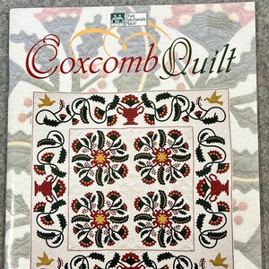 May include: A book cover titled "Coxcomb Quilt" featuring a detailed quilt design. The quilt design has a white background with red, yellow, and black floral patterns. The cover also includes the author's name, Donna Hanson Eines.
