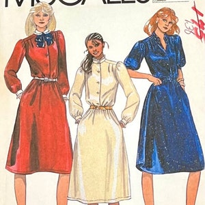 May include: A vintage sewing pattern featuring three different dress styles. The dresses are a red dress with long sleeves and a bow, a cream dress with long sleeves and a collar, and a blue dress with short sleeves and a collar. The pattern is labeled "McCall's 921".