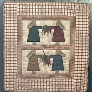 May include: A quilted wall hanging with a brown and white checkered border. The center features four brown, green, red, and blue fabric figures with a heart design and a bunch of red peppers.