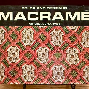 May include: A hardcover book titled Color and Design in Macramé by Virginia I. Harvey. The cover features a detailed, repeating geometric pattern of knotted cord in shades of cream, coral pink, and olive green against a dark background.