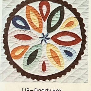 May include: A colorful quilt block with a brown scalloped border. The block features a circular design with a star in the center and radiating petals in shades of red, orange, yellow, green, and blue.