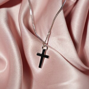 May include: A silver chain necklace with a simple silver cross pendant. The necklace is laying on a pink satin fabric.