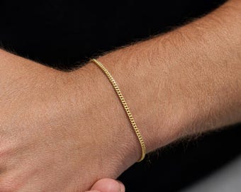 May include: A delicate gold chain bracelet. The bracelet is a thin, continuous strand of small, interlocking links. The bracelet is worn on a wrist, with the skin tone appearing light. The background is dark.