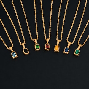 Può includere: Sette collane a catena ritorta color oro, ognuna con un ciondolo rettangolare con una pietra preziosa di colore diverso. Le pietre includono azzurro, nero, verde scuro, rosso, ambra, blu scuro e verde brillante, incastonate in metallo dorato.